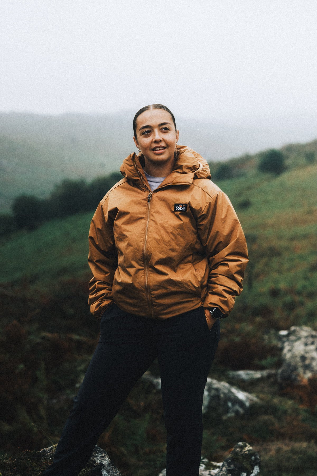 Woman wearing an orange LODE insulated jacket with a raised hood, showcasing its fit, warmth and full-length front zip against a misty mountain backdrop.
