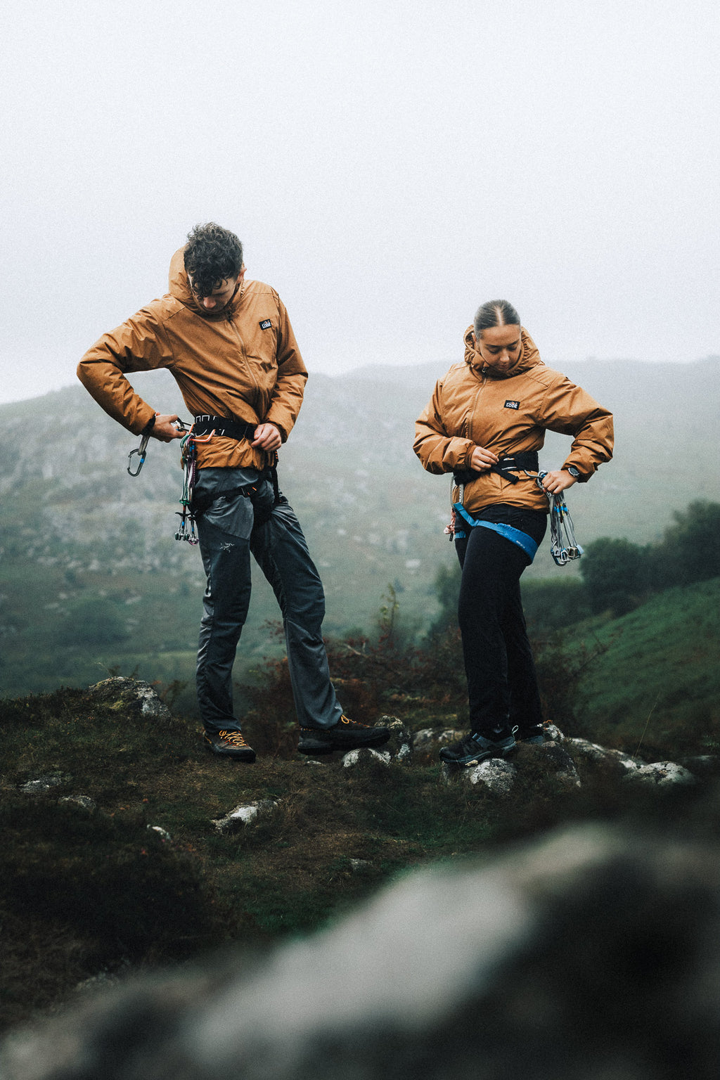 Two climbers wearing orange insulated jackets while adjusting their climbing harnesses on a misty mountainside.