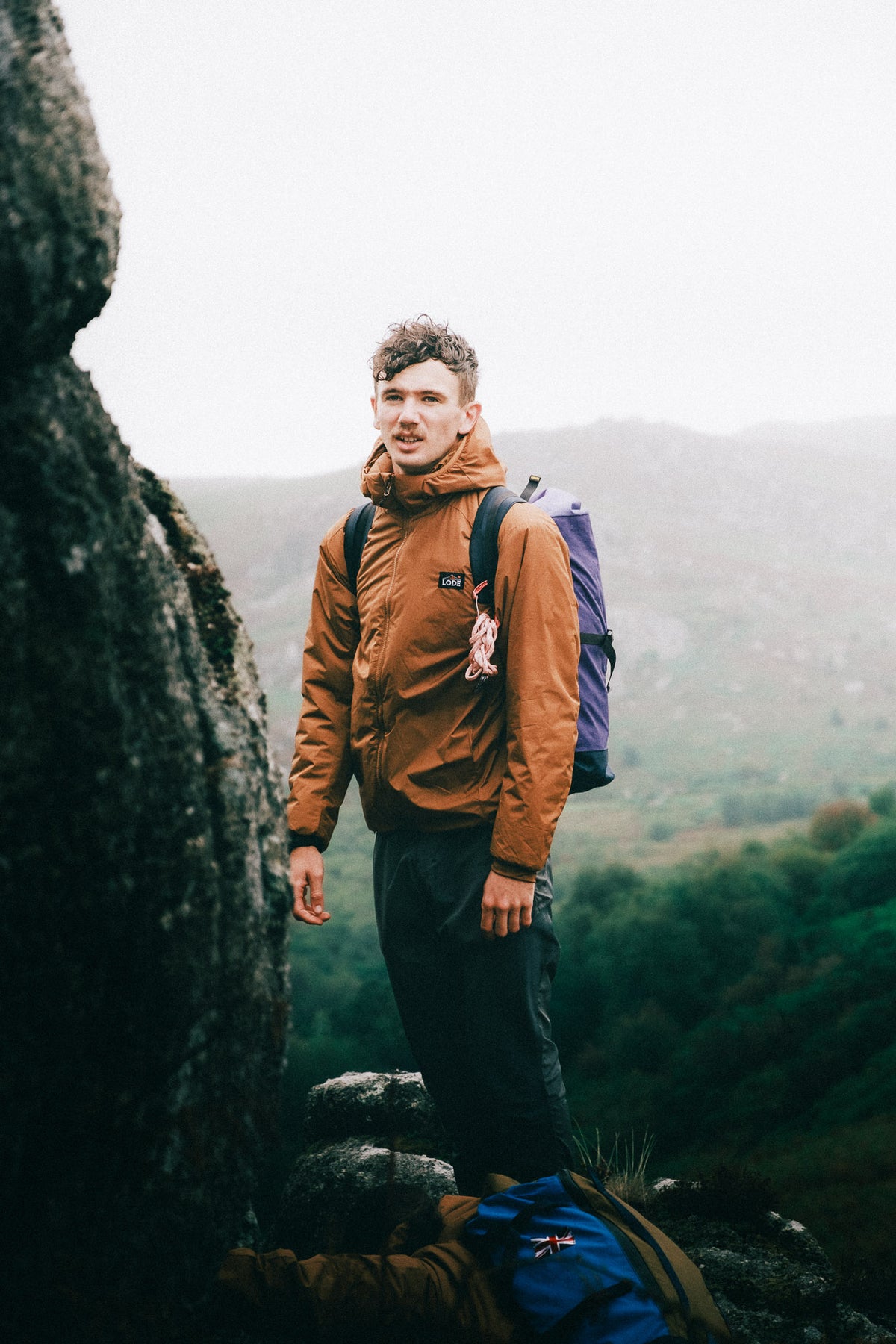 Climber wearing an orange insulated Zawn jacket and carrying a backpack on a rocky mountainside, showcasing its fit and freedom of movement for mountaineering.