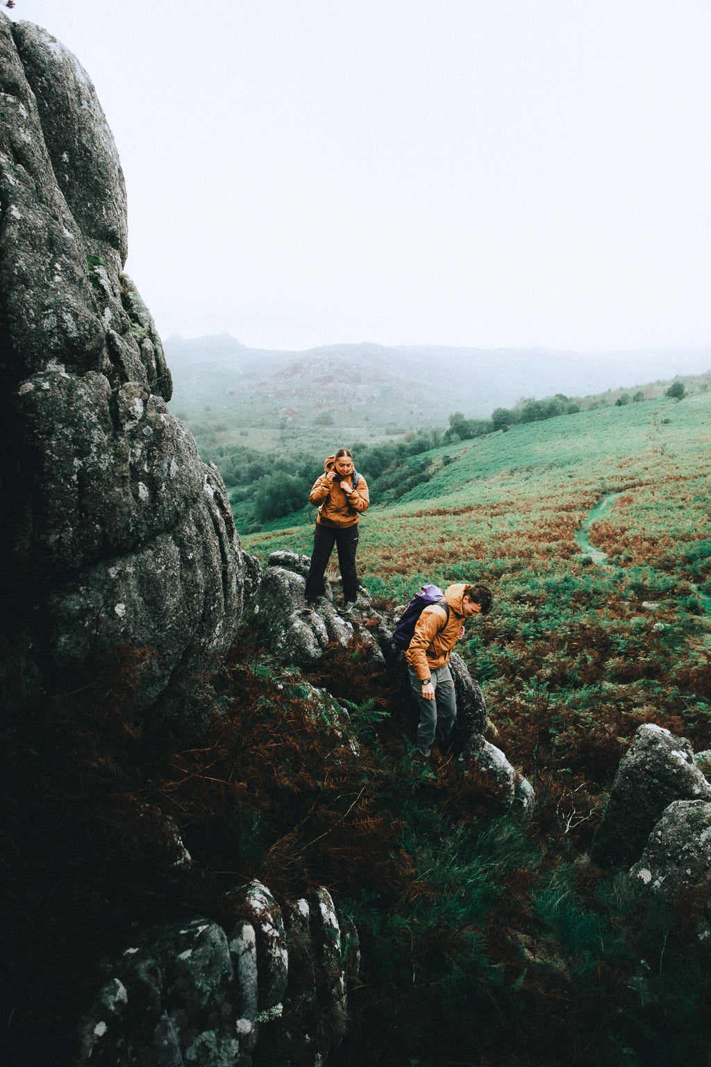 Two climbers wearing orange LODE Zawn insulated jackets while scrambling over rocky terrain on a misty hillside.